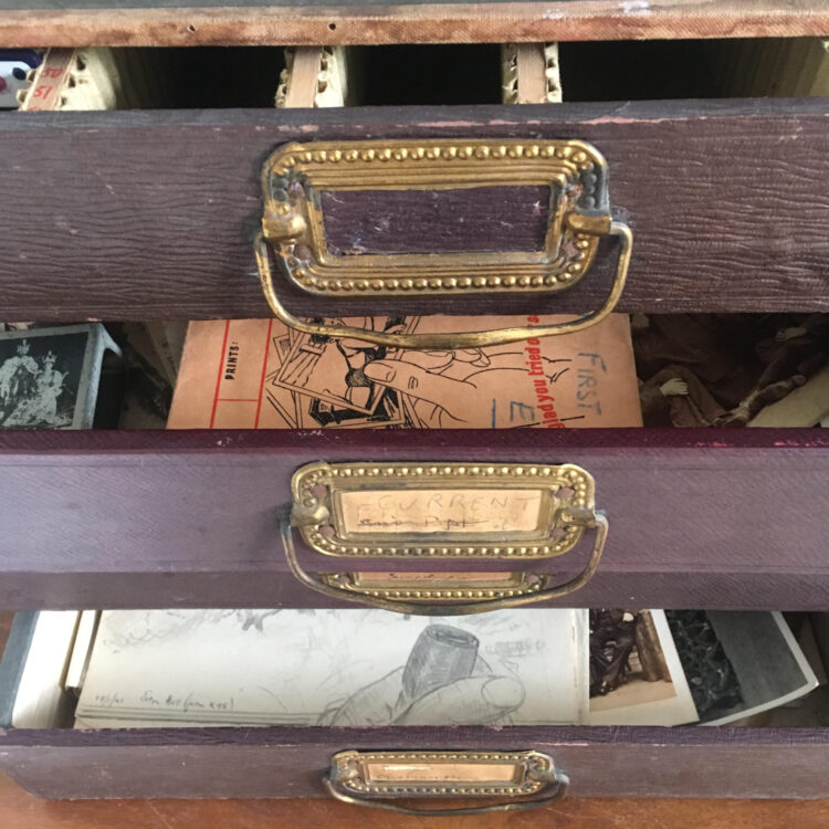 Cabinet with open drawers revealing objects including a 1930's Kodak negative wallet and a drawing of a smoker's pipe in a man's hand 